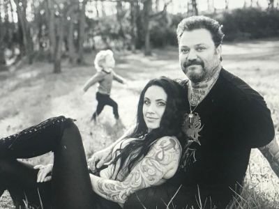 Nicole Boyd is resting on Bam Margera as they are sitting on grass while son, Phoenix Wolf Margera runs in the background.